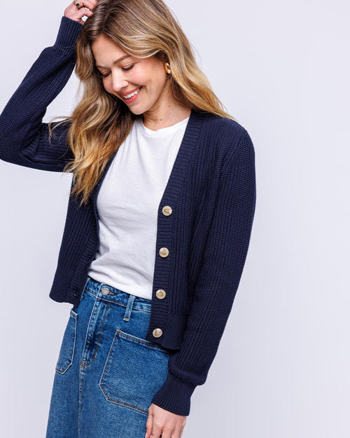 LIFESTYLE / EDITORIAL: Woman wearing navy textured cardigan over white tee and blue jeans, smiling with hand in hair