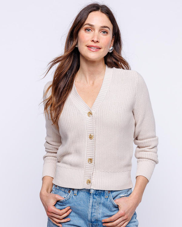 LIFESTYLE / EDITORIAL: Woman wearing oatmeal textured cardigan and blue jeans, standing with hands in pockets against white