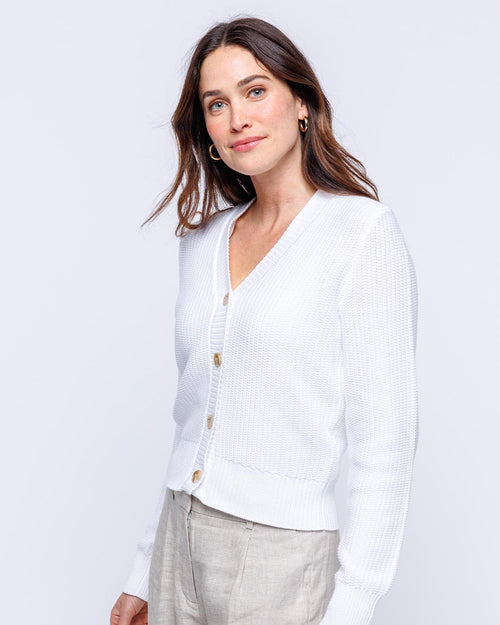 Woman wearing white textured cotton cardigan with buttons, beige pants, standing against light background
