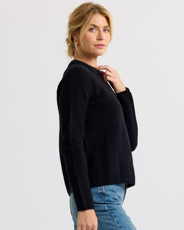 LIFESTYLE / EDITORIAL: Woman wearing black ribbed cashmere sweater and blue jeans, posing sideways against plain background