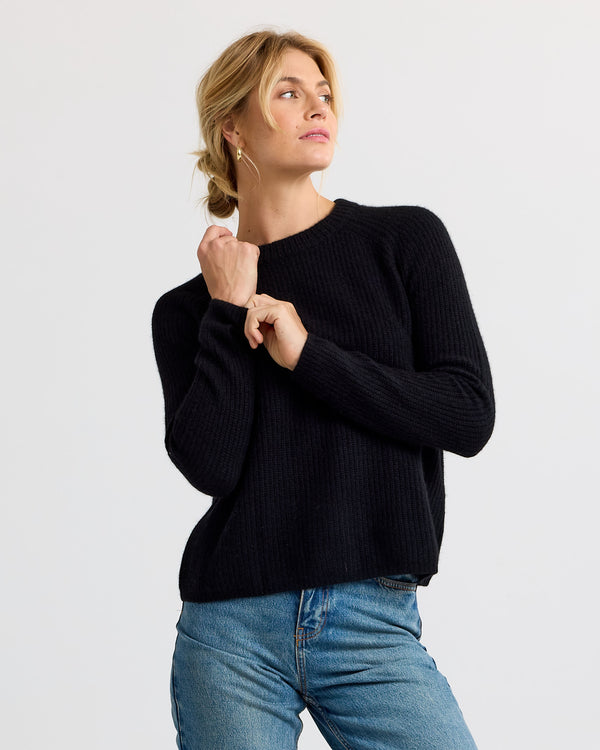 LIFESTYLE / EDITORIAL: Woman wearing black ribbed cashmere sweater and blue jeans, posing against plain background