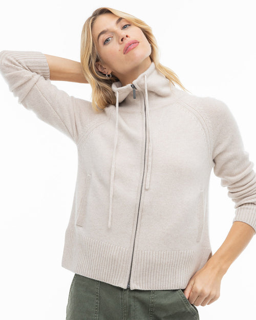 LIFESTYLE / EDITORIAL: Woman wearing oatmeal zip-up cashmere sweater with high collar and drawstrings, posing with hand