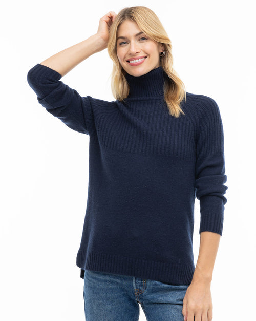 LIFESTYLE / EDITORIAL: Woman in navy turtleneck cashmere sweater and jeans, smiling with hand on head against white