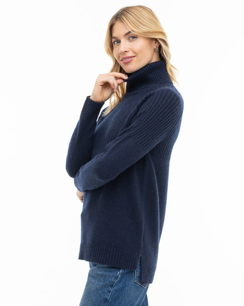 Woman in navy blue turtleneck cashmere sweater and jeans, posing sideways against white background