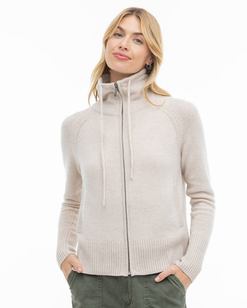Front-facing image of a woman wearing an oatmeal zip-up cashmere sweater with hands in pockets, white background