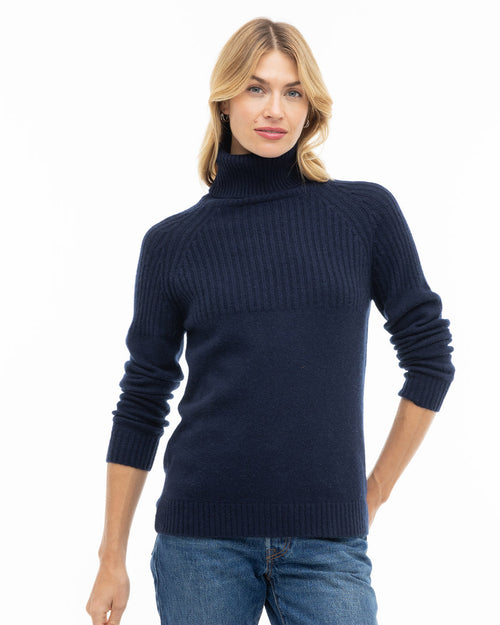 Woman wearing navy blue ribbed turtleneck cashmere sweater and blue jeans, standing against white background