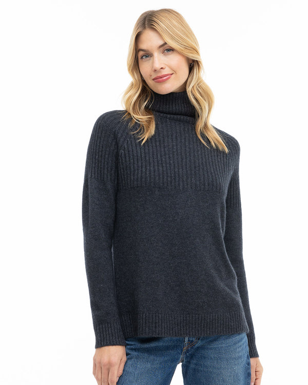 Three-quarter view image of a woman wearing a charcoal ribbed turtleneck cashmere sweater with blue jeans