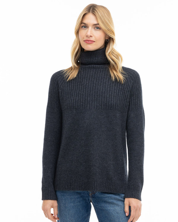 LIFESTYLE / EDITORIAL: Woman wearing charcoal ribbed turtleneck cashmere sweater and blue jeans, standing against white