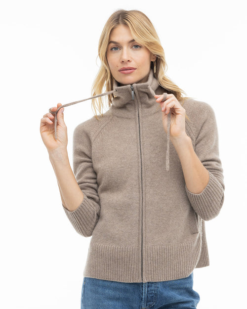 LIFESTYLE / EDITORIAL: Woman wearing a taupe zip-up cashmere sweater with high collar and blue jeans, holding drawstrings