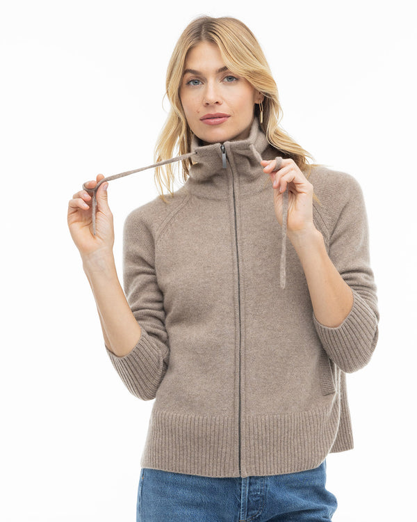LIFESTYLE / EDITORIAL: Woman wearing a taupe zip-up cashmere sweater with high collar and blue jeans, holding drawstrings