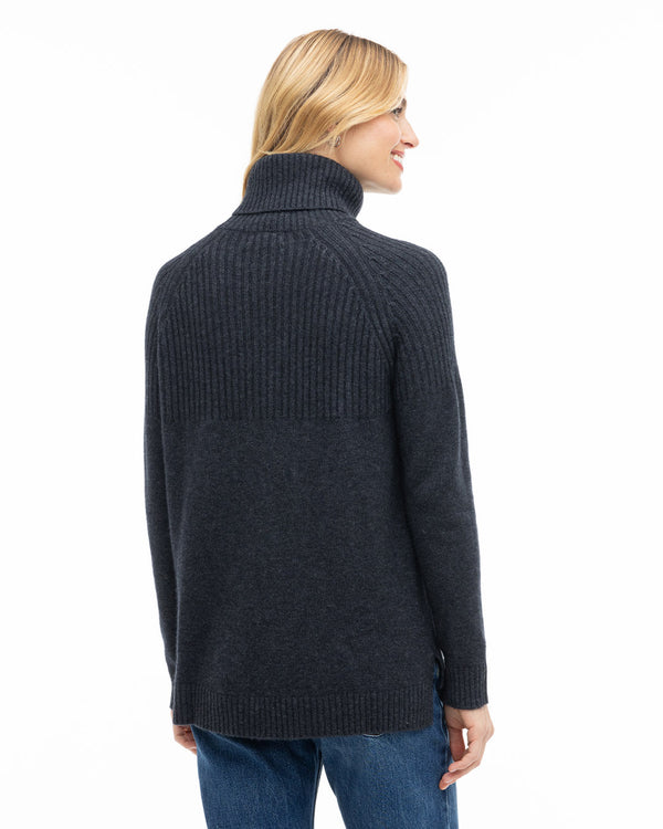 Rear three-quarter view image of a woman wearing a charcoal cashmere turtleneck sweater with blue jeans