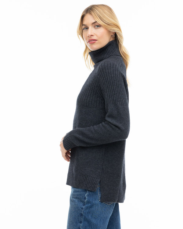 Side profile image of a woman wearing a charcoal ribbed cashmere turtleneck sweater with blue jeans against white background