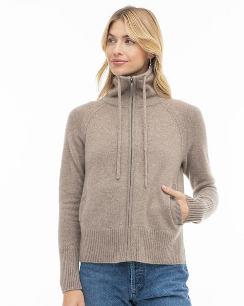 LIFESTYLE / EDITORIAL: Woman wearing driftwood-colored zip-up cashmere sweater with high collar and blue jeans against white