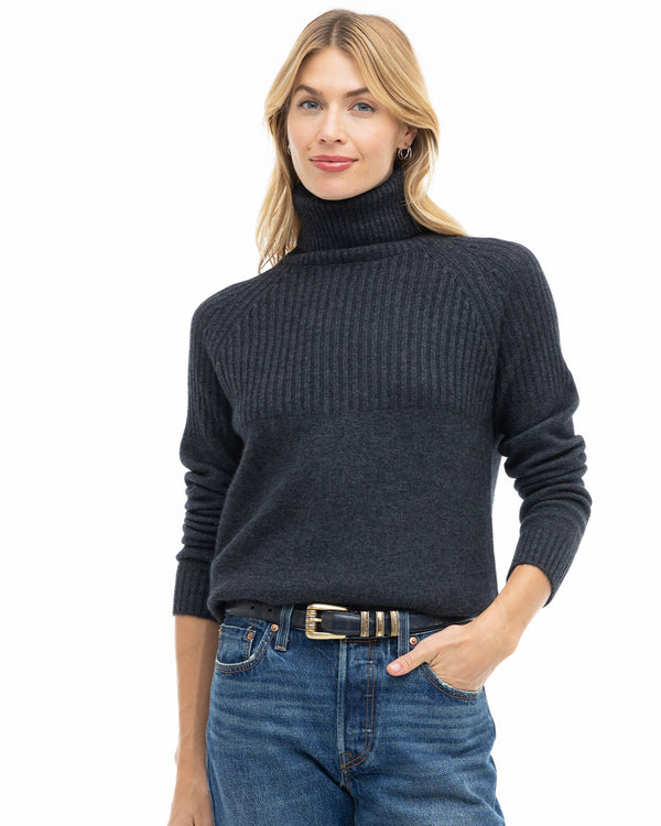 LIFESTYLE / EDITORIAL: Woman wearing charcoal ribbed turtleneck cashmere sweater and blue jeans, hand in pocket, white