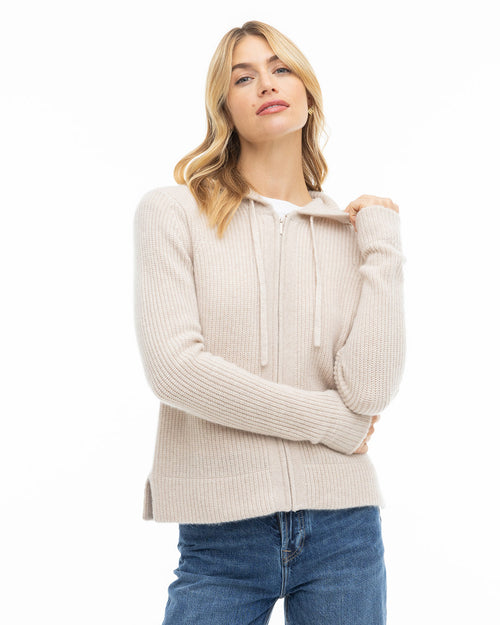 LIFESTYLE / EDITORIAL: Woman wearing oatmeal ribbed zip-up cashmere sweater with hood and blue jeans against white background