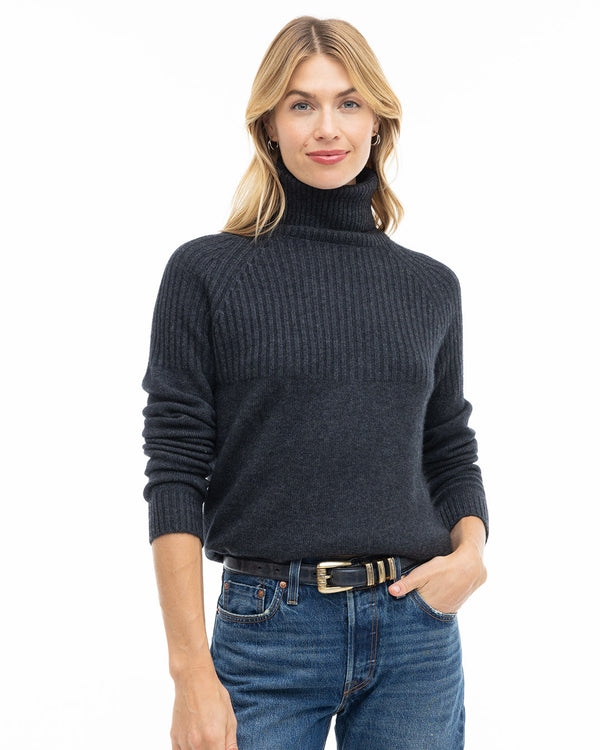 LIFESTYLE / EDITORIAL: Woman wearing charcoal turtleneck cashmere sweater and blue jeans, standing against white background
