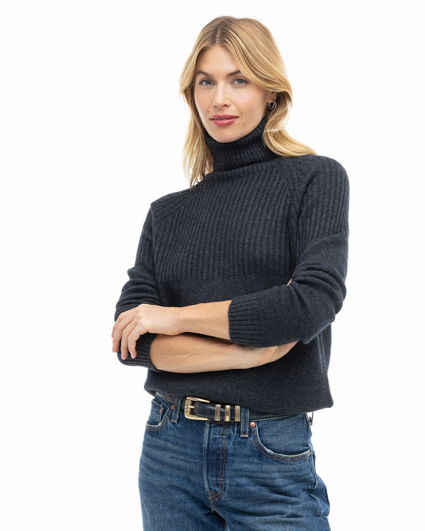 LIFESTYLE / EDITORIAL: Woman wearing charcoal ribbed turtleneck cashmere sweater and blue jeans with black belt, white