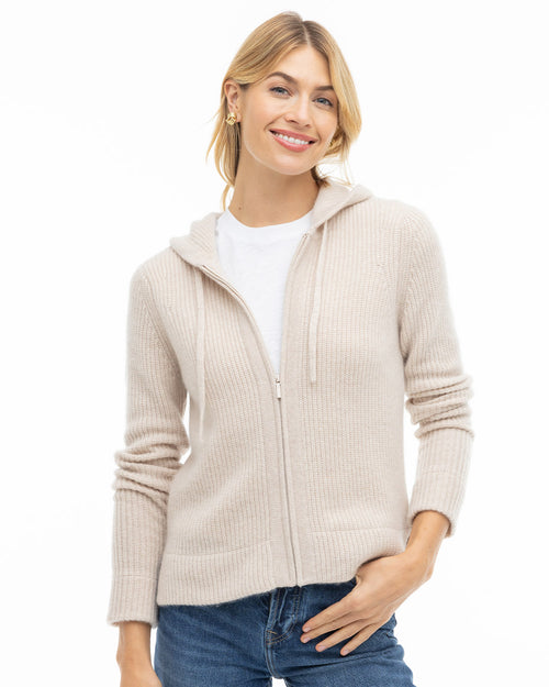 LIFESTYLE / EDITORIAL: Woman wearing oatmeal zip-up cashmere hoodie over white tee and blue jeans, smiling against white