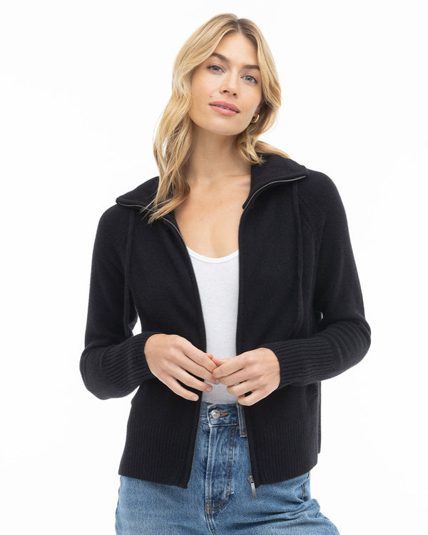Woman wearing black zip-up cashmere sweater over white top and blue jeans, standing against white background