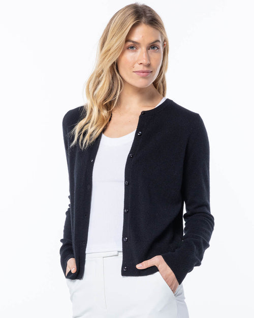Woman wearing black buttoned cashmere cardigan over white top and white pants, standing against white background