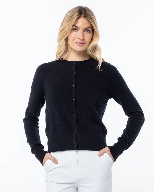 Woman wearing black buttoned cashmere cardigan and white pants, standing with hands in pockets against white background