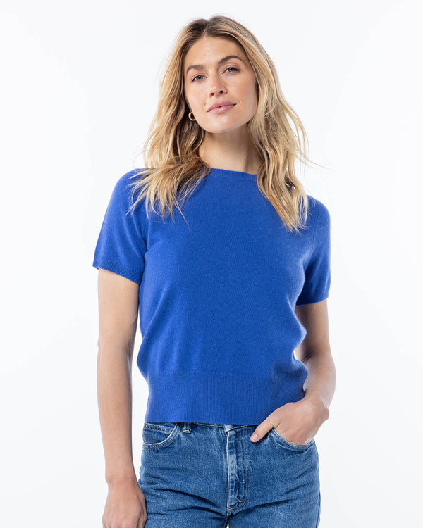 Woman wearing a short-sleeve French blue cashmere sweater and blue jeans, standing against a white background