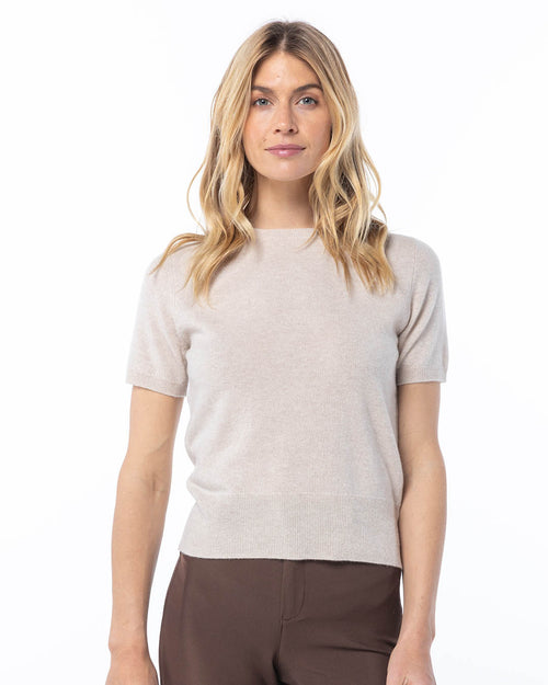 LIFESTYLE / EDITORIAL: Woman wearing oatmeal short-sleeve cashmere sweater and brown pants, standing against white background