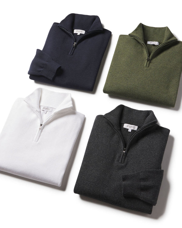 LIFESTYLE / EDITORIAL: Four folded quarter-zip cashmere sweaters in navy, olive, white, and charcoal on white background