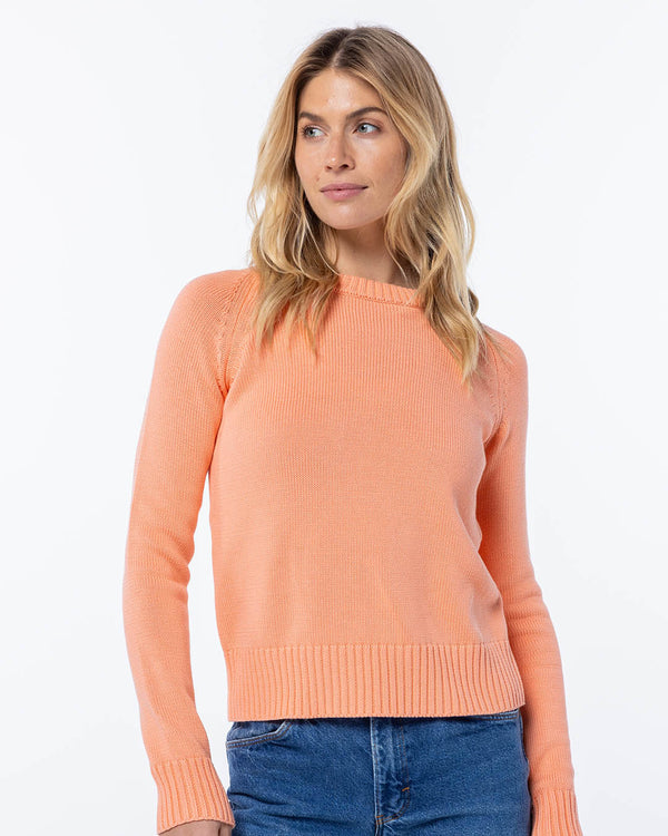 Woman wearing coral cashmere sweater with ribbed cuffs and hem, paired with blue jeans, against white background