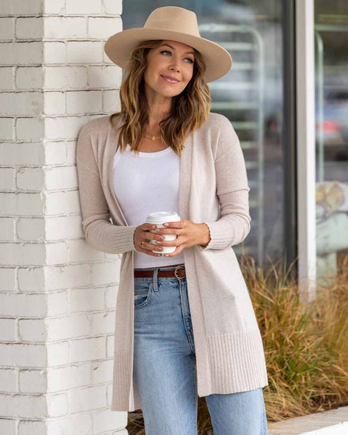 LIFESTYLE / EDITORIAL: Woman in oatmeal long cashmere cardigan, white top, jeans, and hat holding a coffee cup outdoors