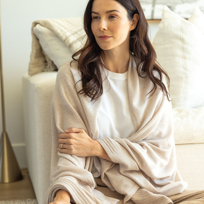 Woman seated on sofa wearing beige cashmere wrap and white shirt, neutral-toned cozy living room background
