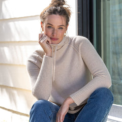 Woman sitting by window wearing beige cashmere turtleneck sweater and blue jeans, natural light background