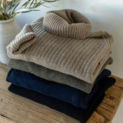 Stack of folded ribbed cashmere sweaters in beige, olive, navy, and black on wooden surface next to a vase with greenery