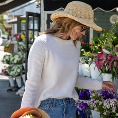 Woman wearing white cashmere sweater and straw hat, standing by flowers at outdoor market