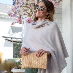 Woman wearing light gray cashmere poncho and sunglasses holding woven clutch outdoors near pink blossoms
