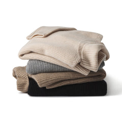 Stack of folded cashmere fisherman turtleneck sweaters in beige, gray, brown, and black on white background