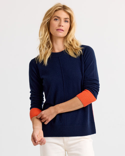 LIFESTYLE / EDITORIAL: Woman wearing navy blue cashmere sweater with orange cuffs and white pants against white background