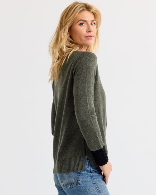 Woman wearing dark olive and black cashmere sweater, shown from side and back, paired with blue jeans against white