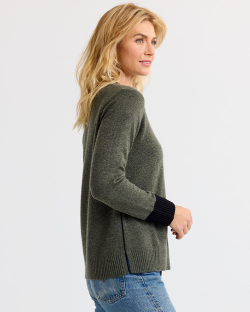 LIFESTYLE / EDITORIAL: Woman in dark olive cashmere sweater with black cuffs, side view, wearing blue jeans against white