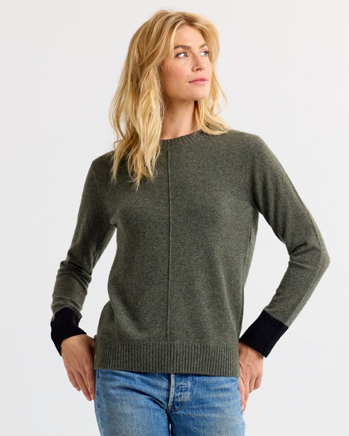 LIFESTYLE / EDITORIAL: Woman wearing dark olive and black cashmere sweater with blue jeans, standing against white background