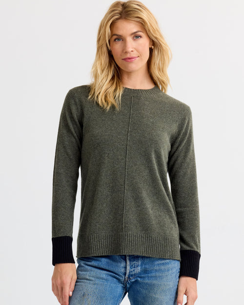 Woman wearing dark olive cashmere sweater with black cuffs and blue jeans against plain white background