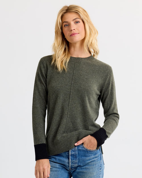 Front-facing image of a woman wearing dark olive cashmere sweater with black cuffs and blue jeans against white background