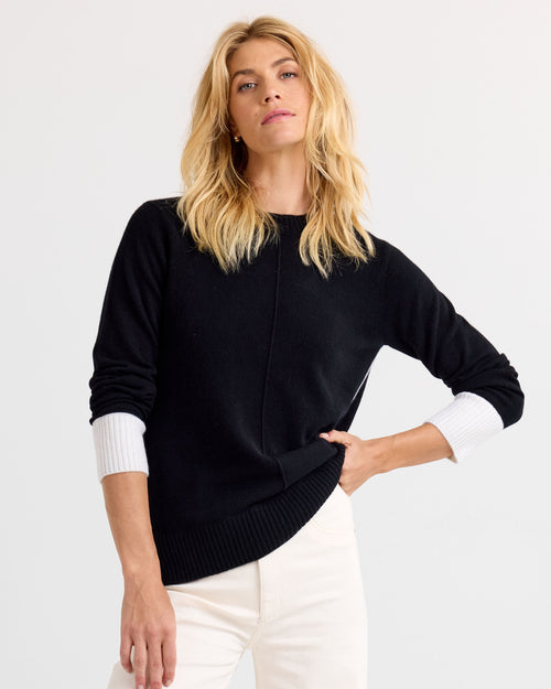 Three-quarter view image of a woman wearing a black cashmere sweater with white cuffs and white pants, hand on hip.