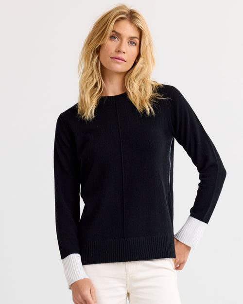 LIFESTYLE / EDITORIAL: Woman wearing black cashmere sweater with white cuffs and white pants against plain background