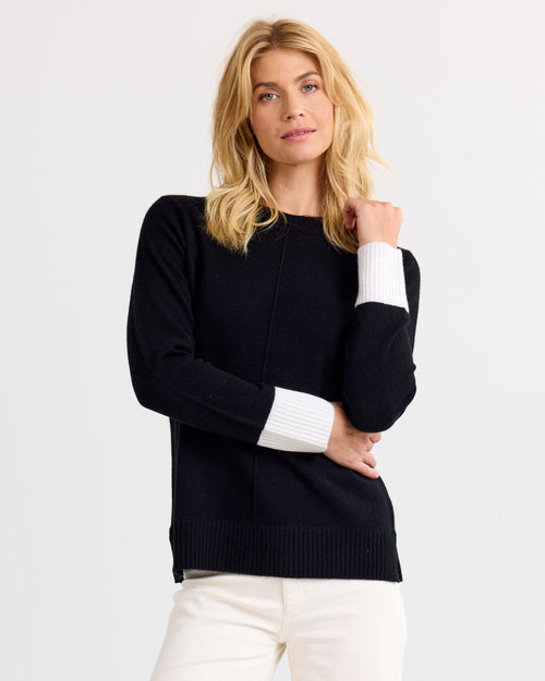 Three-quarter view image of a woman wearing a black cashmere sweater with white cuffs and white pants against plain