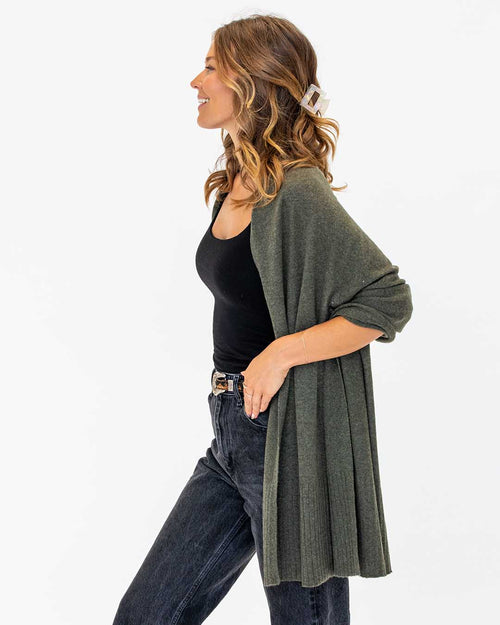 LIFESTYLE / EDITORIAL: Woman in dark olive cashmere wrap, black tank top, and dark jeans, standing in profile against white