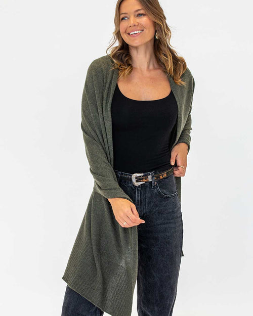 LIFESTYLE / EDITORIAL: Woman wearing dark olive cashmere wrap over black top and dark jeans, smiling against white background