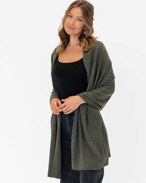 Woman wearing dark olive cashmere travel wrap over black top and dark jeans against white background