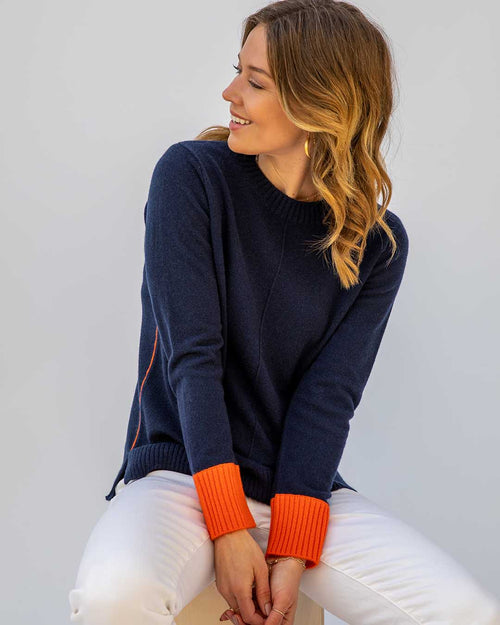Woman wearing navy cashmere sweater with orange cuffs and white pants, sitting and looking to the side