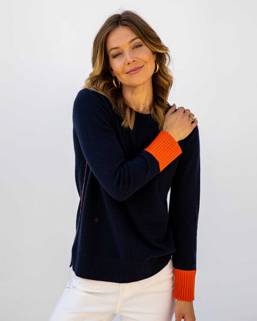 Woman wearing navy cashmere sweater with orange cuffs and white pants against plain background